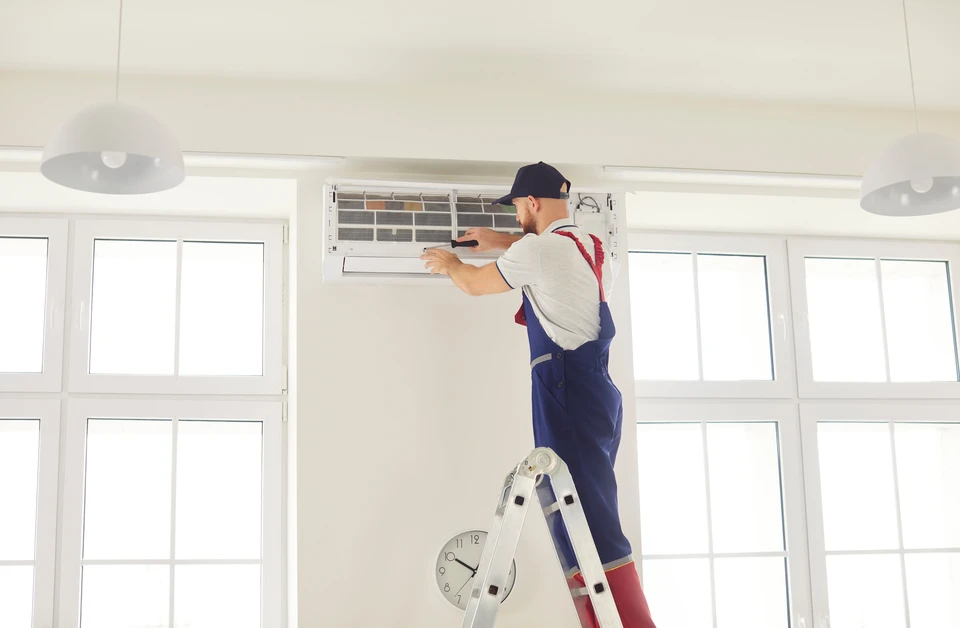 Technicien en climatisation travaillant dans une pi&egrave;ce r&eacute;sidentielle, portant une combinaison rouge et gants bleus, manipulant des outils pr&egrave;s d'une unit&eacute; de climatisation murale ouverte